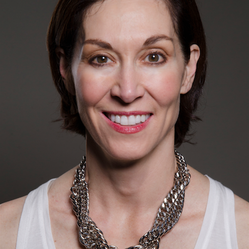A woman with short dark brown hair smiles at the camera. She is wearing a sleeveless white top and a thick silver braided necklace, with a dark neutral background.
